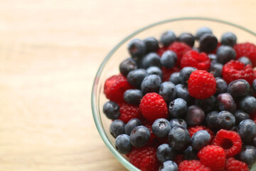 Glass bowl filled with raspberries and blueberries on wooden table. Selective focus.