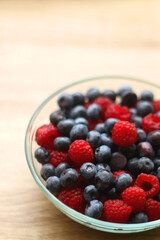 Glass bowl filled with raspberries and blueberries on wooden table. Selective focus.