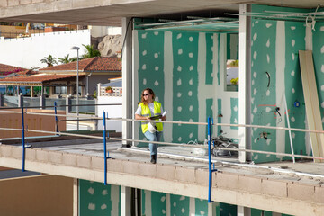 Female architect or supervisor standing on an unfinished balcony of a modern building under construction, wearing a high-visibility vest and safety boots while holding rolled-up blueprints and tablet