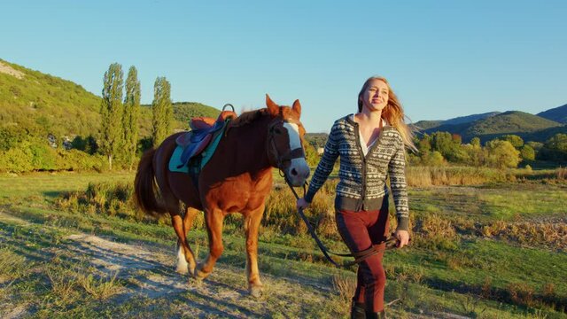 European long-haired girl leads chestnut horse by reins along hill. Panorama, landscape with mountain, wooded steppe. Young woman going with palfrey in countryside. Horsewoman walks bay stallion
