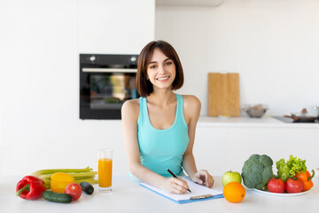 Happy fitness lady in sportswear writing daily ration diet or healthy recipe, standing in kitchen