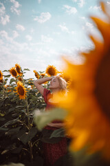 Adorable young caucasian girl posing in a sunflower field in the sun light. Woman in red summer style dress with open hands over cloudy blue sky