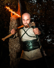 A stern bearded bald Viking with an axe and a torch in the night forest