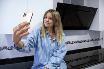 Mujer sentada en la cocina de su casa. Mujer vestida con camisa ancha. Chica con el móvil haciéndose selfies y sonriendo. 