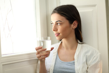 Woman holding water glass lokking through window at home