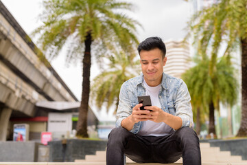 Portrait of handsome young man sitting outdoors in city during summer while using mobile phone