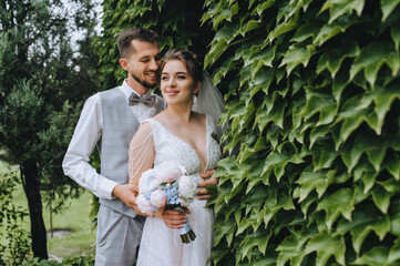 A beautiful, sweet bride in a white dress with a bouquet and a stylish groom in a gray tuxedo are hugging against the background of green foliage of decorative grapes. Wedding photography.