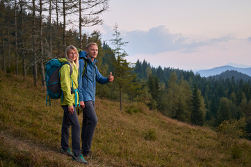 Smiling husband and wife with backpacks standing on grassy downhill trail against the backdrop of mountain forests. Positive male while trekking adventure showing cool gesture.