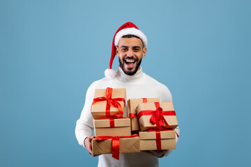Buying holiday gifts. Overjoyed arab man in santa hat holding stack of present boxes, smiling over blue background