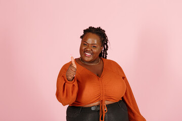 Smiling young black plus size body positive woman with dreadlocks in orange top shows thumb-up standing on pink background in studio closeup