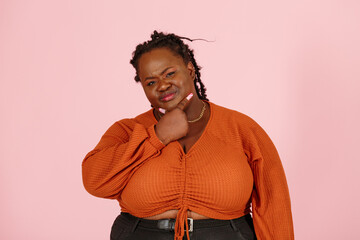 Skeptical young black plus size body positive woman with dreadlocks in orange top touches chin looking at camera on light pink background in studio closeup