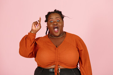 Funny surprised young black overweight body positive woman with dreadlocks in orange top gets cool idea looking at camera on light pink background in studio closeup - 466753476