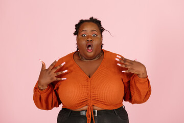 Amazed young black plus size body positive woman with dreadlocks in orange top throws up hands on light pink background in studio closeup