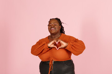 Smiling young black plus size body positive woman with dreadlocks in orange top shows heart with palms on light pink background studio portrait close view