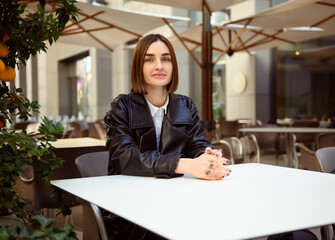 Attractive confident beautiful brunette young woman with short haircut in a black eco leather jacket sitting at a table on the terrace of a cozy cafe, confidently looking at the camera