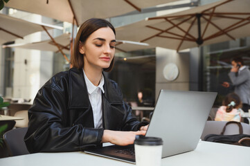 Naklejka na ściany i meble Confident young business woman, copywriter, developer, freelancer, journalist working remotely on laptop, writing text, enjoying distant work in outside summer cafeteria on beautiful early spring day