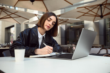 Young beautiful Caucasian woman, copywriter writing on notepad, using laptop sitting at the table...