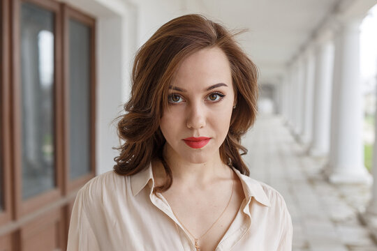 closeup portrait of pretty caucasian young woman in beige blouse, seductive red lipstick and wavy hair standing on veranda of cafe. white concrete pillars and lanterns in background