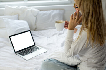 Blonde woman is sitting on the white bed and looks at the mockup laptop. Remote work. Covid 19. Young woman in on bed using laptop With blank mockup template on screen display. Work from home. White