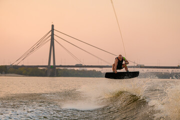 active man holds rope and masterfully jumps on wakeboard on city bridge background.
