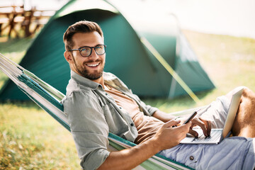 Happy man uses smartphone and laptop while camping in nature and looking at camera.