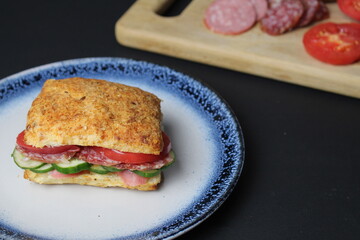 sandwich sandwich of white square bun with vegetables tomato cucumber sausage ham on a blue plate. in the background is a tray board with ingredients  vegetables tomatoes on a black background. 