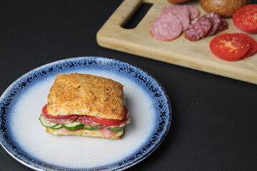 sandwich sandwich of white square bun with vegetables tomato cucumber sausage ham on a blue plate. in the background is a tray board with ingredients vegetables tomatoes on a black background. 