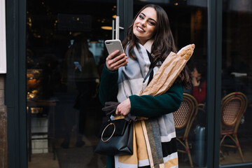 Happy female client of bakery near entrance with smartphone and purchase
