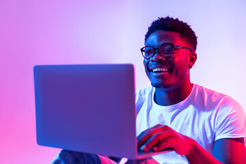 Happy young black man using laptop computer for online work, education or communication in neon light - obrazy, fototapety, plakaty