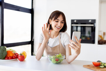 Video blog about healthy food. Happy young woman with bowl of fresh salad on table recording video on smartphone