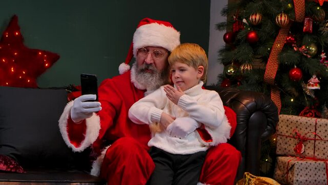 Santa Claus with a child sitting on his lap are taking a selfie using a smartphone. Concept of the New Year spirit, holidays and celebrations. Close up. Slow motion.