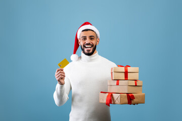 Christmas gifts shopping. Happy arab guy holding credit card and pile of wrapped present boxes,...