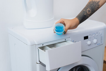 cropped view of tattooed woman holding cap with liquid detergent near washing machine.