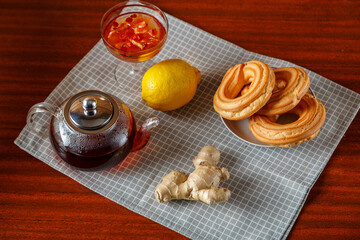A kettle of strong hot tea on a napkin next to lemon, ginger and jam and donuts.