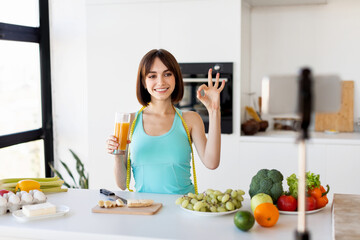 Happy fit woman drinking fresh detox and talking at smartphone camera, showing ok gesture, shooting video for vlog