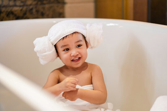 Asian Cute Baby Girl In A Bathrobe And Hair Wrapped In Towel Having Fun In A Bathtub