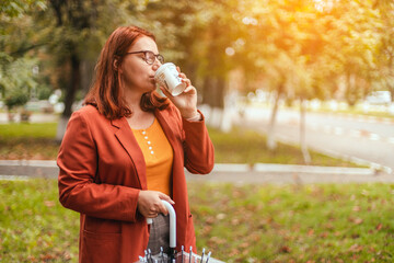 Beautiful young woman having fun drinking morning coffee in sunlight while walking through autumn...