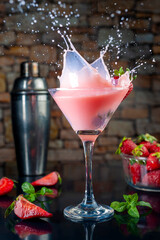 cocktail glass with a shot of strawberries and natural strawberries, metal shaker, forming a splash on black table and stone wall