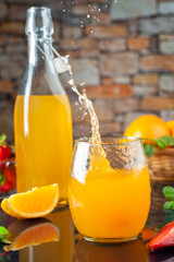 natural orange juice with whole oranges and strawberries with glass and bottle filled with liquid flying splash on black background and stone wall