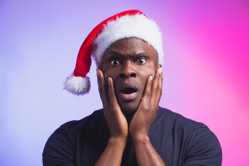 Portrait of surprised african american man in santa hat and casual t-shirt on colourful background, christmas time. Winter holidays and emotional people concept.