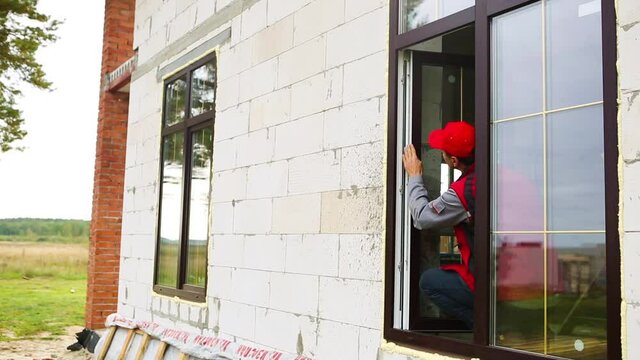 The window installer adjusts the hinges and checks new windows in the cottage under construction. Ordering windows with golden layout and brown lamination for a private house. A worker in uniform
