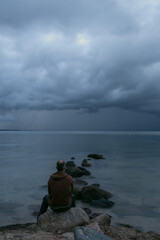 Fototapeta premium Mann sitzt alleine auf einem Stein am Meer mit dunklen Regenwolken am Himmel.