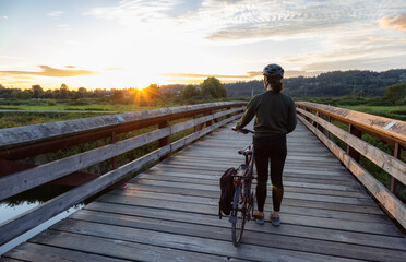 White Caucasian Woman with a bicycle on a wooden bridge in a modern city park during summer sunset. Port Coquitlam, Vancouver, British Columbia, Canada.
