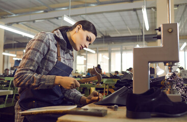 Female worker using professional tools while making new leather boots. Beautiful young woman working with hammer and nail at shoe factory workshop. Manufacturing industry, footwear production concept