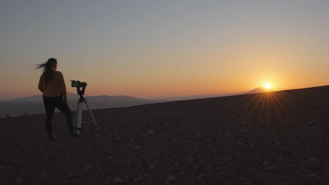 Wide view of a woman filming the sunset with camera and tripod in the desert. Slow motion