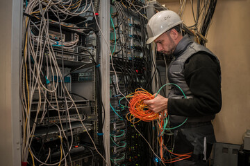 An engineer in a white helmet works in a server room. Technician connects fiber optic cables with...