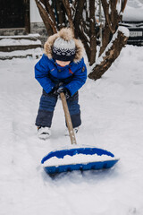 Little boy with a shovel in hand remove snow in backyard, snow removal. Kid in blue jacket cleans snow with shovel after snowfall
