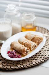 pancakes, rolled into rolls, on a white plate with frozen raspberries and honey with wooden forks on the background of a glass of milk, jars of sugar and honey on a wicker napkin