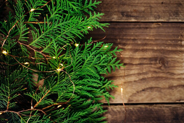 Background of Christmas lights of green thuja branches. A festive shiny garland is intertwined with branches on the brown surface of wooden boards.