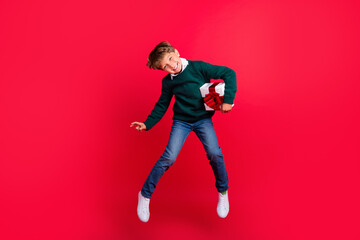 Fototapeta na wymiar Photo of funny cute schoolboy dressed green sweater jumping high holding gift box smiling isolated red color background